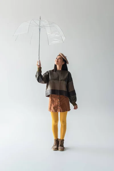 Full length view of asian woman in stylish clothes smiling under umbrella on grey — Stock Photo