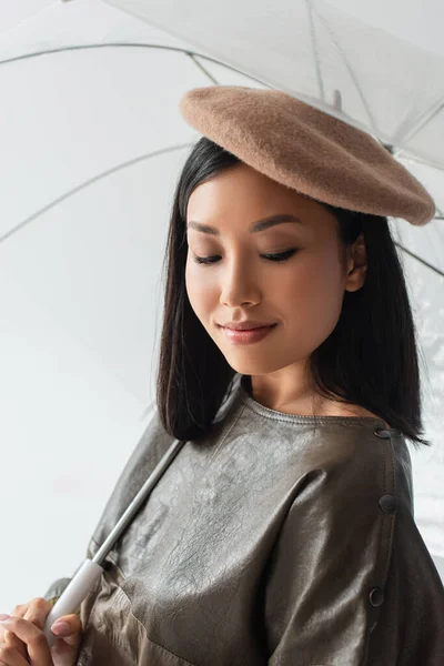 Красивая азиатка в бежевом берете улыбается под прозрачным зонтиком, изолированным на сером — стоковое фото