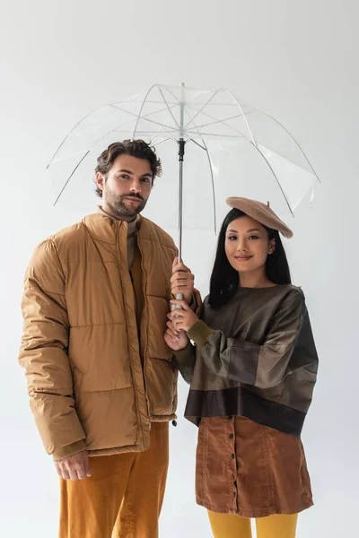 Man in beige puffer jacket and asian woman in leather pullover looking at camera under umbrella isolated on grey — Stock Photo