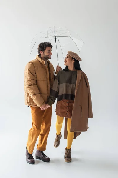 Full length view of pleased multiethnic couple holding hands under transparent umbrella on grey — Stock Photo