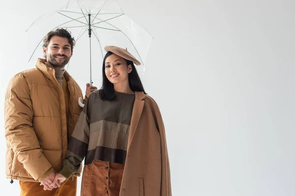 Межрасовая пара в стильной одежде, держась за руки и улыбаясь под прозрачным зонтиком, изолированным на сером — стоковое фото
