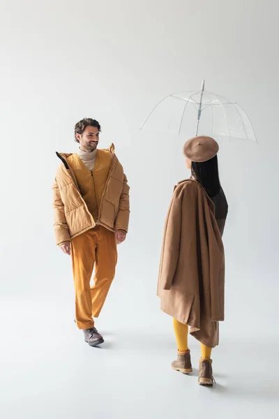 Femme en parapluie transparent et homme souriant en veste de bouffée beige marchant les uns vers les autres sur gris — Photo de stock