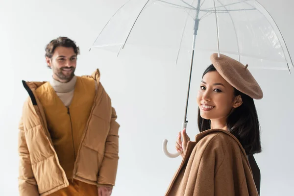 Азиатка с прозрачным зонтиком, улыбающаяся в камеру рядом с размытым мужчиной, изолированным на сером — стоковое фото
