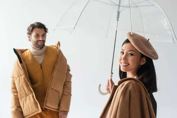 Asiático mujer con transparente paraguas sonriendo en cámara cerca borrosa hombre aislado en gris - foto de stock