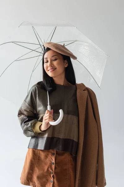 Trendy asian woman standing under transparent umbrella and smiling with closed eyes isolated on grey — Stock Photo