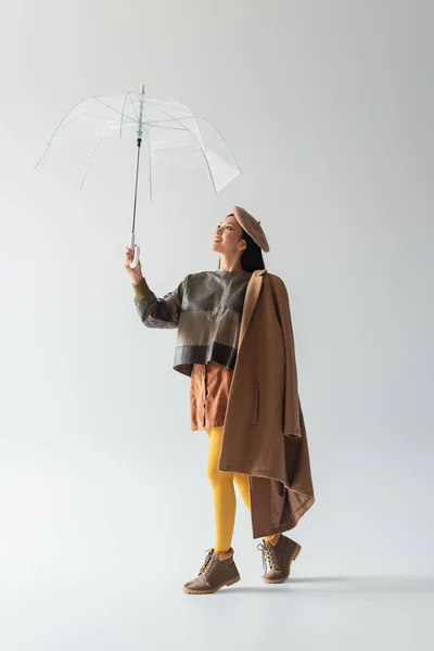 Vista completa de mujer asiática en jersey de cuero y medias amarillas bajo paraguas transparente en gris - foto de stock