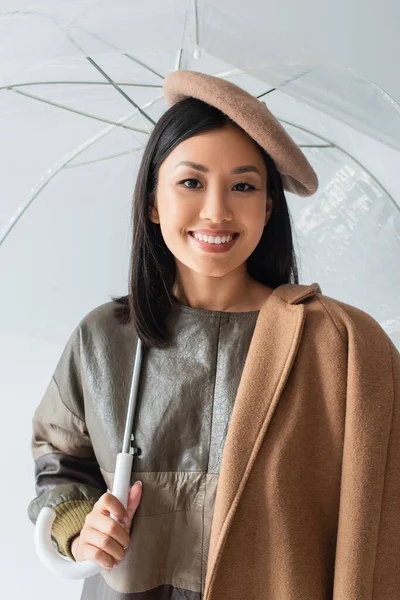 Happy asian woman in beret looking at camera under transparent umbrella isolated on grey — Stock Photo