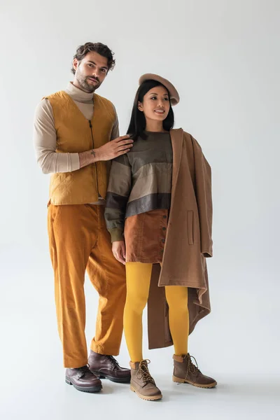 Hombre de moda mirando a la cámara cerca de mujer asiática en boina y medias amarillas en gris - foto de stock