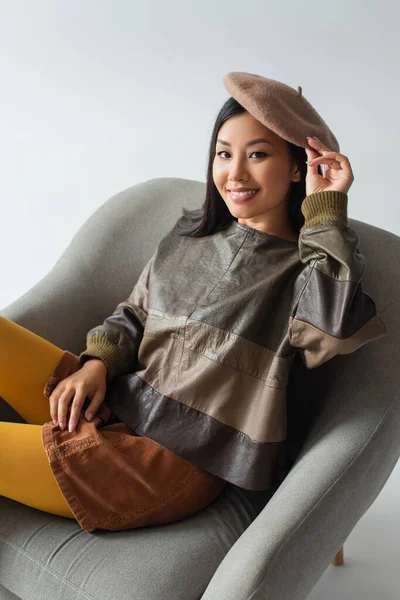 Ásia mulher no couro jaqueta e boina sentado no poltrona no cinza — Fotografia de Stock