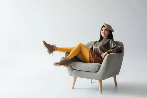 Vista completa de mujer asiática feliz en jersey de cuero y medias amarillas sentadas en sillón en gris - foto de stock