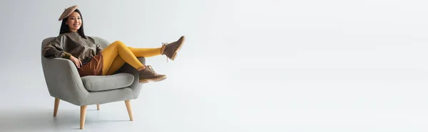 Volle Länge Ansicht der fröhlichen und modischen asiatischen Frau posiert im Sessel auf grau, Banner — Stockfoto