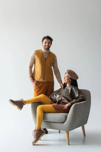 Hombre feliz y elegante de pie con la mano en el bolsillo cerca de la mujer asiática en sillón en gris - foto de stock