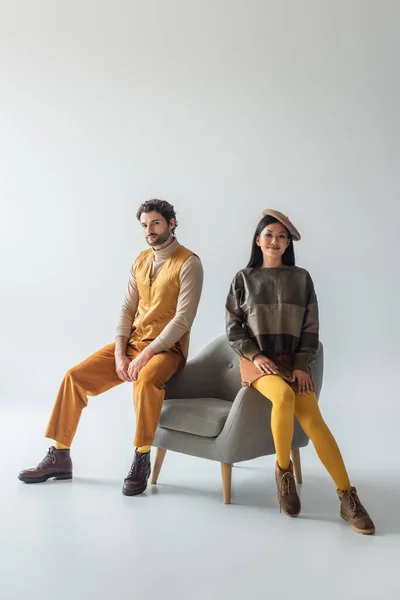 Multiethnic couple in fashionable clothes sitting on armchair and looking at camera on grey — Stock Photo