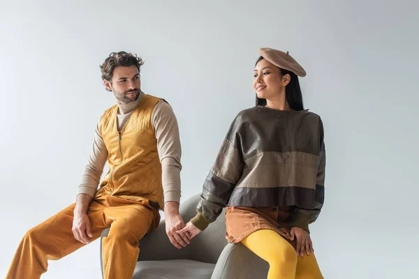 Donna asiatica sorridente in berretto e pullover di pelle tenendosi per mano con uomo in giubbotto giallo isolato su grigio — Foto stock