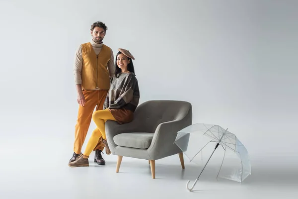 Cheerful asian woman sitting on armchair near trendy man and transparent umbrella on grey — Stock Photo