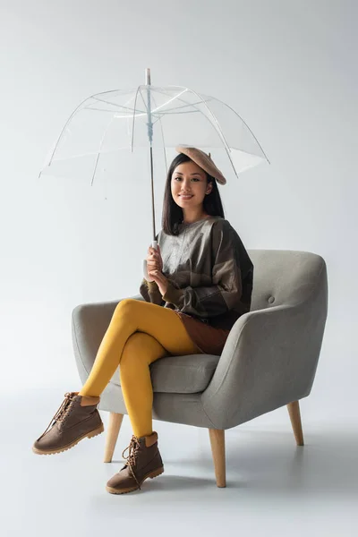 Mujer asiática sonriente en jersey de cuero y medias amarillas sentadas en sillón bajo paraguas transparente sobre gris - foto de stock