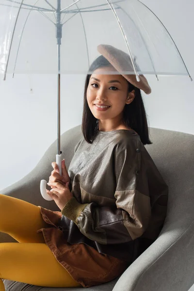 Стильная азиатка, сидящая в кресле под зонтиком и улыбающаяся в камеру, изолированную на сером — стоковое фото