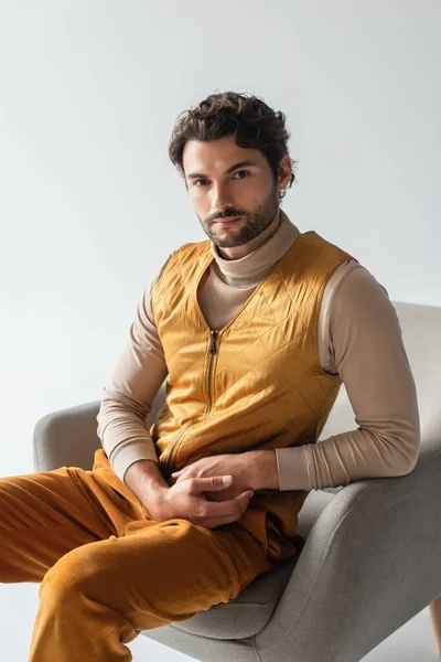 Stylish man in yellow vest and trousers sitting in armchair and looking at camera isolated on grey — Stock Photo