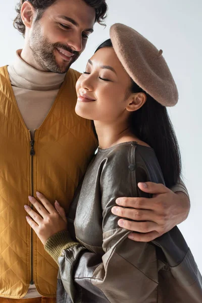 Feliz y elegante hombre abrazando asiático mujer sonriendo con cerrado ojos aislado en gris - foto de stock