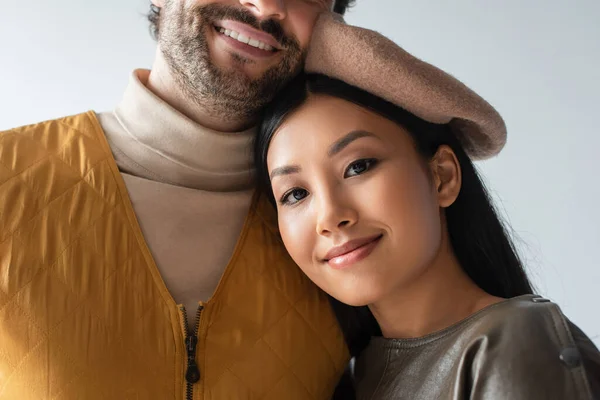 Счастливая азиатская женщина в берете смотрит в камеру, опираясь на улыбающегося мужчину, изолированного на сером — стоковое фото
