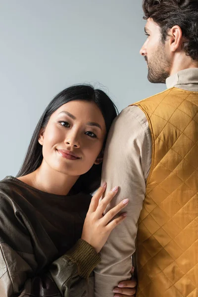 Rückansicht eines Mannes in gelber Weste neben einer asiatischen Frau im Lederpullover, die isoliert auf grau in die Kamera lächelt — Stockfoto