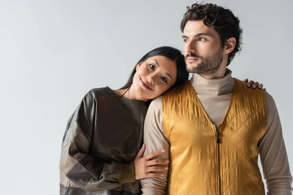 Feliz asiática mujer apoyada en morena hombre en amarillo chaleco aislado en gris - foto de stock