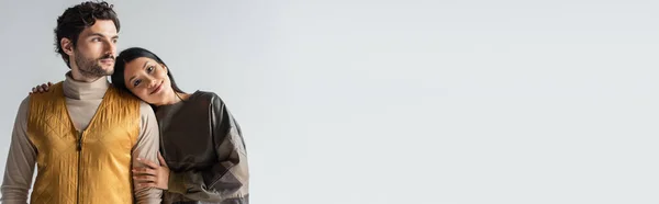 Heureux asiatique femme en cuir pull regarder caméra près de branché homme isolé sur gris, bannière — Photo de stock