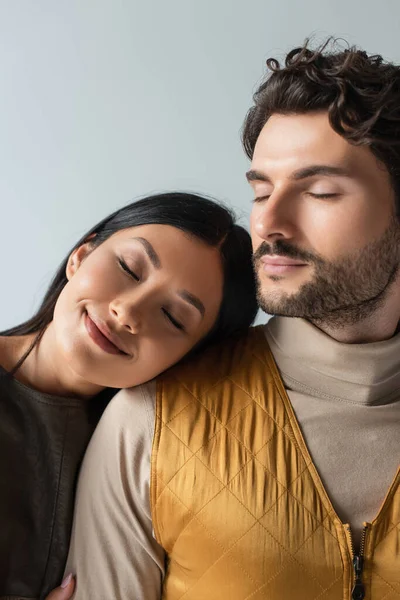 Lächelnde asiatische Frau mit geschlossenen Augen stützt sich auf stilvolle brünette Mann isoliert auf grau — Stockfoto