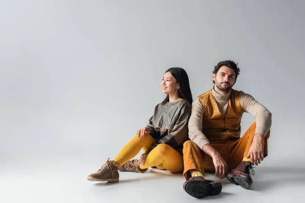 Uomo alla moda sorridente alla fotocamera mentre seduto vicino felice donna asiatica sul grigio — Foto stock