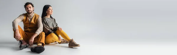 Fashionable man sitting near smiling asian woman and looking at camera on grey, banner — Stock Photo