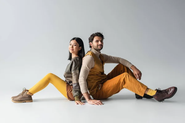 Pareja interracial elegante sonriendo a la cámara mientras se sienta espalda con espalda en gris - foto de stock