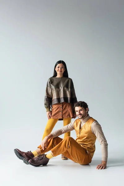 Smiling man in yellow trousers sitting near asian woman in leather jacket on grey — Stock Photo