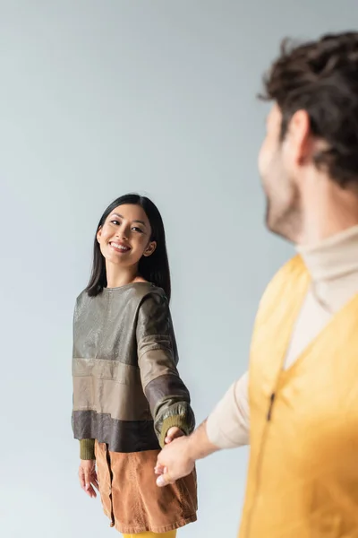 Весела азіатська жінка в шкіряному пуловері тримає руки з розмитим чоловіком ізольовано на сірому — стокове фото