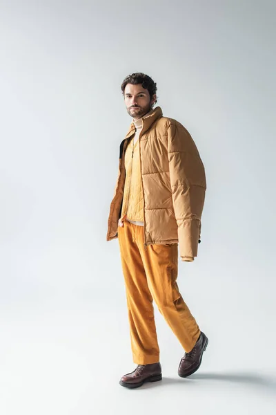 Full length view of man in warm puffer jacket and yellow trousers posing with hand in pocket on grey — Stock Photo