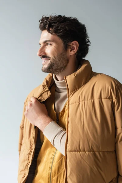 Hombre feliz y elegante en chaqueta hinchable caliente mirando hacia otro lado aislado en gris - foto de stock