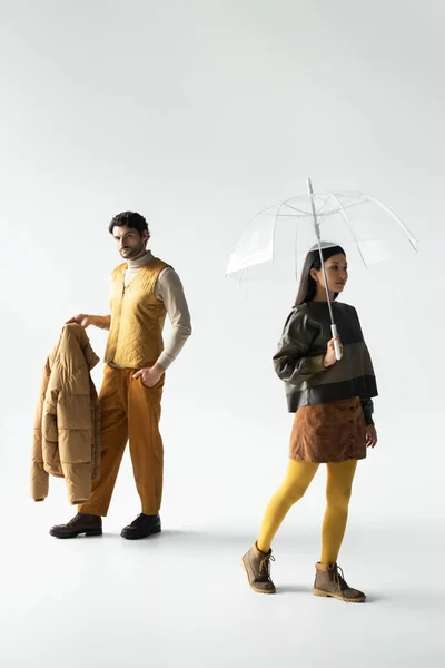 Mann mit Kugeljacke und asiatische Frau mit durchsichtigem Regenschirm gehen auf grau — Stockfoto