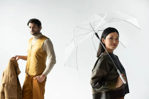 Asian woman smiling at camera under transparent umbrella near man holding puffer jacket on grey — Stock Photo