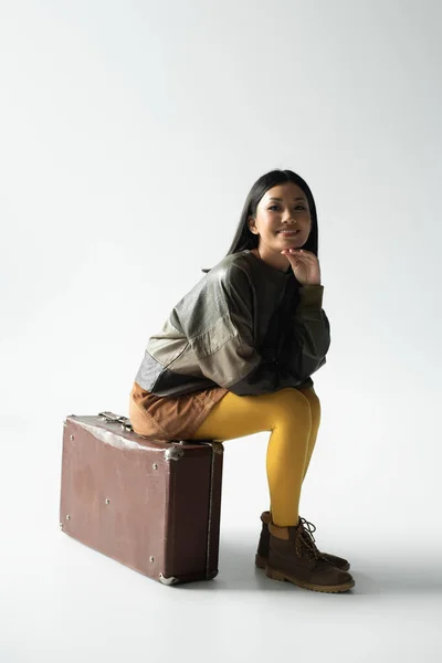 Asian woman in trendy clothing sitting on vintage suitcase on grey — Stock Photo