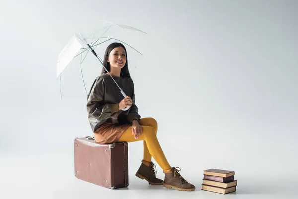 Glückliche asiatische Frau mit transparentem Regenschirm sitzt auf Vintage-Koffer neben Büchern auf grau — Stockfoto