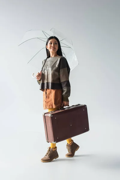 Vue pleine longueur de femme asiatique élégante avec valise vintage marchant sous parapluie transparent sur gris — Photo de stock