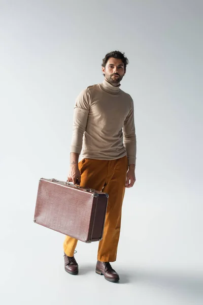 Полный вид человека в бежевой водолазке и желтых брюках, идущего с винтажным чемоданом на сером — стоковое фото
