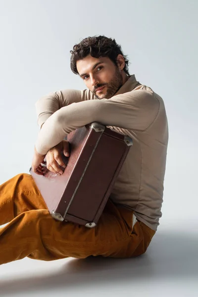 Trendy man in yellow trousers and beige turtleneck sitting with vintage suitcase on grey — Stock Photo