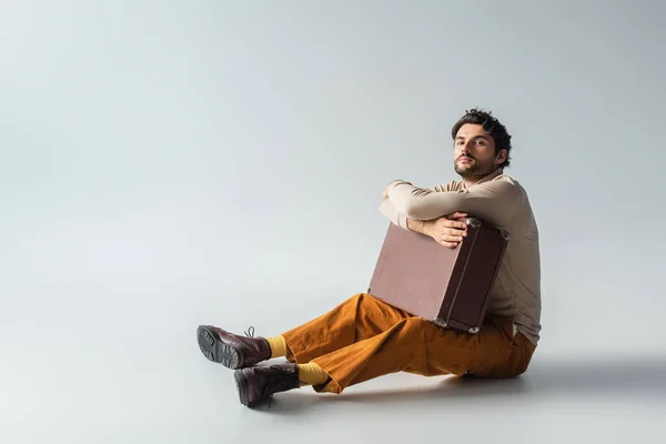 Vue pleine longueur de l'homme en vêtements à la mode assis avec valise vintage sur gris — Photo de stock