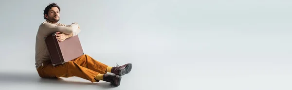 Vista completa del hombre en pantalones amarillos y botas marrones sentados con maleta retro en gris, bandera - foto de stock