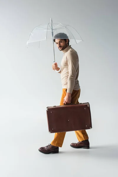 Modischer Mann mit Vintage-Koffer spaziert unter transparentem Regenschirm auf grau — Stockfoto