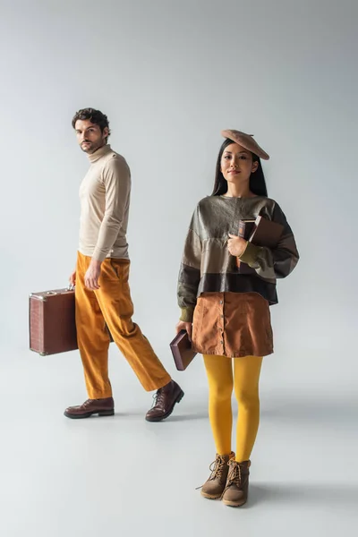 Vista completa de hombre con maleta vintage y mujer asiática con libros en gris - foto de stock