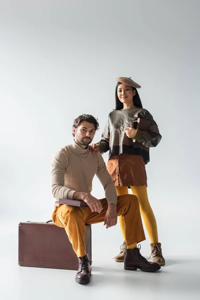 Stylish asian woman holding books near man sitting on vintage suitcase on grey — Stock Photo