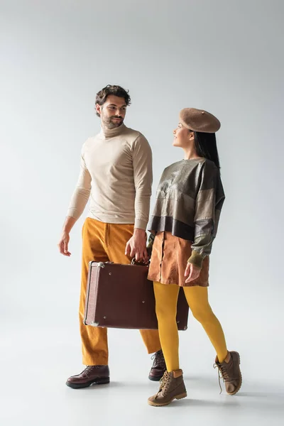 Модная межрасовая пара, несущая винтажный чемодан и улыбающаяся друг другу на сером — стоковое фото
