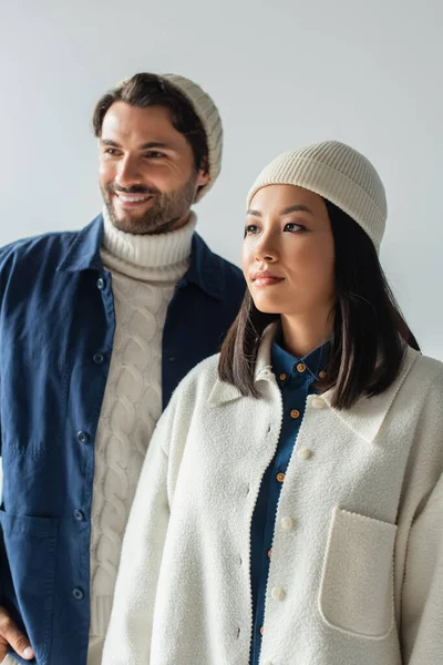 Mujer asiática en chaqueta blanca y gorro cerca de hombre de moda sonriendo sobre fondo borroso aislado en gris - foto de stock