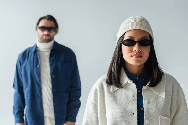 Junge asiatische Frau mit dunkler Sonnenbrille und weißer Jacke, die in die Kamera schaut, in der Nähe eines verschwommenen Mannes, isoliert auf grau — Stockfoto